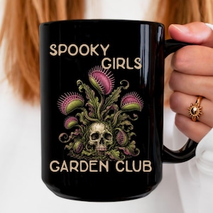 May include: A black ceramic mug featuring a graphic print of a human skull surrounded by green and pink Venus flytrap plants. The text SPOOKY GIRLS GARDEN CLUB is printed in a light tan, sans-serif font above and below the central botanical illustration.