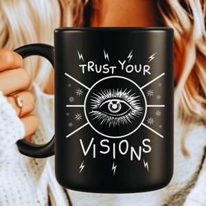 May include: Black ceramic mug with a white graphic of an eye inside a circle, with the words "TRUST YOUR VISIONS" and lightning bolt designs. The mug has a black handle and is held by a person.