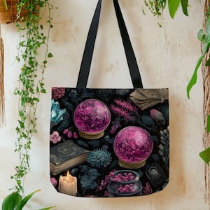 May include: Black tote bag featuring a mystical design with two pink crystal balls, an open book, and a lit candle. The bag has black handles and a dark floral background with accents of blue and pink.