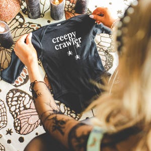 May include: A black baby onesie with the words "creepy crawler" in white script and three hanging spider graphics. The onesie is being held up by a person. The background includes candles and a decorative tablecloth.