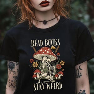 May include: Black t-shirt with a whimsical design featuring a skeleton reading under a mushroom. The design includes the text "READ BOOKS STAY WEIRD" in a decorative font, surrounded by flowers and other mushrooms.