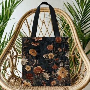 May include: A tote bag with a dark background and a floral design featuring sunflowers and other flowers in shades of orange, yellow, and white. The bag has black handles and is displayed on a wicker chair.