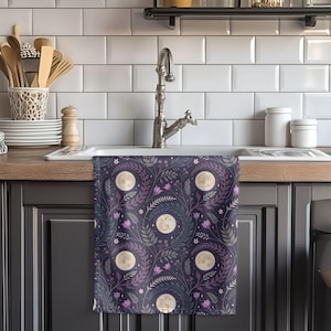 May include: A rectangular kitchen towel featuring a repeating pattern of full moons, purple tulip-like flowers, and dark green leafy branches set against a deep navy blue background. The design includes small white stars scattered throughout the fabric.