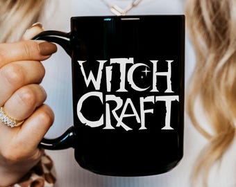 May include: Black ceramic mug with the white text "WITCH CRAFT". The mug has a black handle and is held by a person. The text includes a crescent moon and star design.