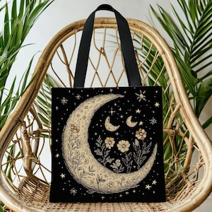 May include: Black tote bag featuring a crescent moon design with flowers and stars. The moon is beige with detailed floral and botanical illustrations. The bag has black handles and is displayed on a wicker chair.