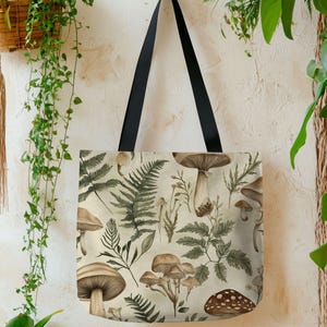 May include: A beige tote bag with a repeating pattern of mushrooms and ferns in shades of brown and green. The bag has black handles and is hanging against a textured, off-white wall with hanging plants.
