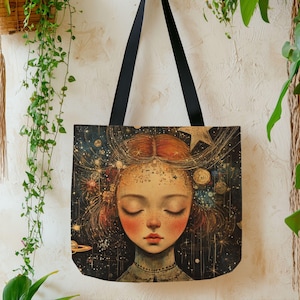 May include: A tote bag with a celestial design. The bag features a portrait of a woman with closed eyes, surrounded by stars and galaxies. The bag has black straps and is made of a durable fabric. The color palette includes deep blues, gold, and red.