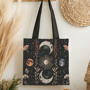 May include: Black tote bag featuring a celestial design with moons, suns, and floral elements in shades of peach, cream, and gold. The bag has black handles and a rectangular shape, perfect for carrying items.