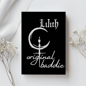 May include: A black rectangular print featuring white text and a graphic. The text reads "Lilith" above a crescent moon and a cross-like symbol, with "original baddie" below. The print is displayed against a white background with floral accents.