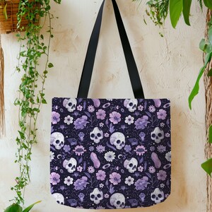 May include: A tote bag with a dark purple background featuring a repeating pattern of white skulls, purple flowers, and other botanical elements. The bag has black handles and is hanging against a neutral wall with plants.