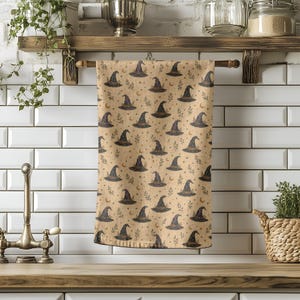 May include: A rectangular kitchen towel featuring a repeating pattern of dark pointed witch hats, small crescent moons, and delicate leafy sprigs printed on a light tan background. The fabric is draped over a horizontal wooden rod.