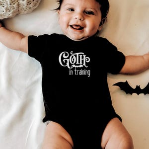 May include: A black baby onesie with the text "Goth in training" in white script. A black bat-shaped decoration is next to the baby. The baby is smiling and lying on a white surface.