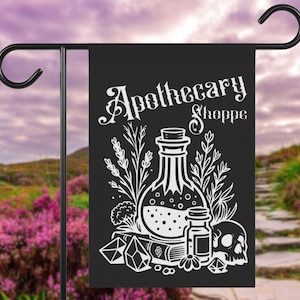 May include: Black garden flag with white illustration and text "Apothecary Shoppe." The design includes a potion bottle, smaller vials, a skull, crystals, and botanical elements. The flag is hanging on a black metal garden stake.