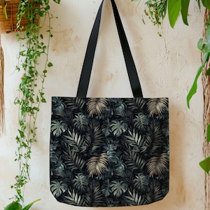 May include: Black tote bag with a tropical leaf pattern in shades of green, gray, and gold. The bag has black straps and is hanging against a neutral-colored wall with hanging plants.