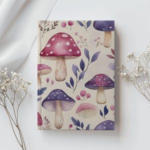 May include: A notebook featuring a whimsical mushroom pattern. The cover displays a repeating design of pink and purple mushrooms with white spots, interspersed with blue leaves and berries, set against a cream background.