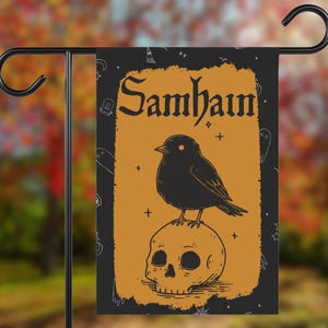 May include: A black garden flag with the word "Samhain" in a decorative font. The flag features a black bird perched on a skull against an orange background with a black border. The flag is displayed on a black metal stand.