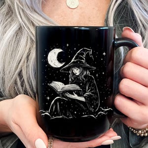 May include: Black ceramic mug featuring a white illustration of a witch reading a book under a crescent moon and stars. The witch wears a pointed hat and robe. The mug has a large handle.