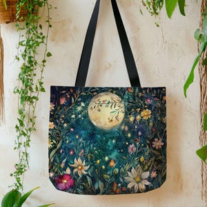 May include: A tote bag with black straps, featuring a whimsical design of a full moon surrounded by colorful flowers and foliage. The artwork has a dark teal background with a starry night sky, creating a dreamy, nature-inspired aesthetic.