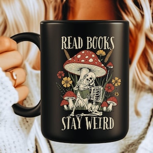 May include: Black ceramic mug with a whimsical design. The mug features the text "READ BOOKS STAY WEIRD" with an illustration of a skeleton reading under a mushroom, surrounded by flowers and other mushrooms. The mug has a black handle.