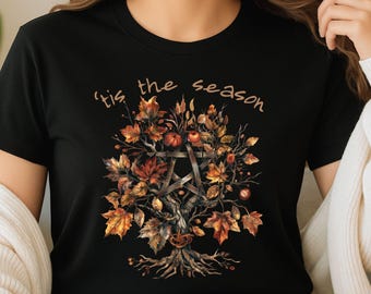 May include: Black t-shirt featuring a fall-themed graphic. The design includes a tree with autumn leaves, a pentagram, and the phrase "'tis the season" in a script font. The leaves are in shades of orange, brown, and yellow.