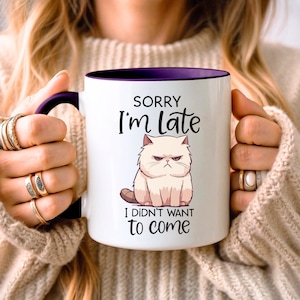 May include: A white ceramic mug with a dark purple interior and handle. The front features a graphic of a grumpy light-colored cat with the text "SORRY I'm Late I DIDN'T WANT to come" printed in black, stylized lettering above and below the cat.