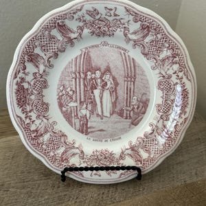May include: A white ceramic plate with a scalloped edge and a maroon transferware print. The center depicts a wedding scene with the text "UN MARIAGE A LA CAMPAGNE" and "LA SORTIE DE L'EGLISE". The border features floral and scroll patterns.