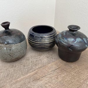 May include: Three ceramic vessels including a round vase with a speckled grey base and dark top, a bowl with horizontal ridges and a dark blue glaze, and a dark, glossy vase with a wide shoulder and a narrow neck, all arranged together.