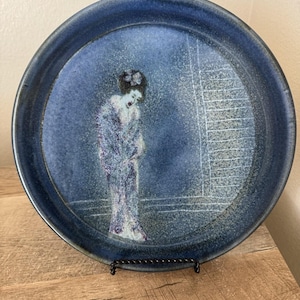 May include: A round ceramic plate with a deep blue, mottled glaze. The center features a stylized illustration of a person with dark hair wearing a long, textured robe. To the right, a series of thin, horizontal white lines form a rectangular pattern.