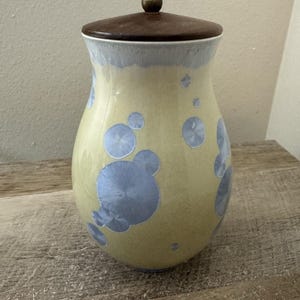May include: A ceramic jar with a pale yellow glaze featuring blue crystalline patterns. The jar has a rounded body and a dark brown wooden lid topped with a small, round, metallic-colored knob. The surface displays a smooth, glossy finish.