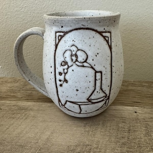 May include: A speckled off-white ceramic mug with a curved handle. The front features a dark brown line drawing of an orchid in a vase, enclosed within a rectangular frame with rounded corners, set against a light-colored, textured clay surface.