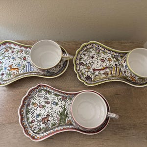 May include: Three ceramic snack sets, each featuring a scalloped-edge tray and a matching cup. The white surfaces are decorated with colorful, intricate hand-painted illustrations of animals, foliage, and architectural elements in various hues.