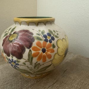 May include: A round ceramic vase with a cream-colored base featuring hand-painted floral motifs in shades of deep purple, bright orange, yellow, and blue, accented by green leaves and a yellow rim with a subtle drip-like glaze pattern.
