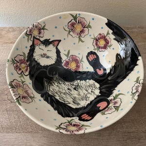 May include: A round ceramic bowl with a cream-colored base featuring a hand-painted illustration of a black and white cat lying on its back. The cat is surrounded by pink flowers with yellow centers and small blue dots scattered across the interior surface.