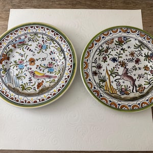 May include: Two round ceramic plates featuring hand-painted designs. The left plate displays a bird, while the right plate shows a deer. Both plates are decorated with intricate floral patterns in blue, yellow, green, and brown, finished with a scalloped border.