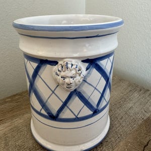 May include: A white ceramic planter featuring a hand-painted blue lattice pattern and a sculpted human face relief on the front. The cylindrical vessel has a wide, flared rim and a smooth, glossy finish, showcasing a classic design aesthetic.