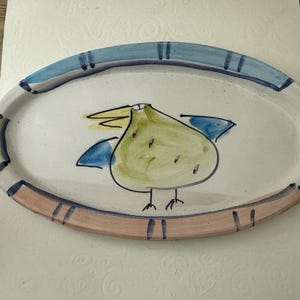 May include: An oval ceramic platter with a cream-colored base featuring a hand-painted green bird with blue wings and a yellow beak. The rim is decorated with alternating segments of blue, yellow, and light pink, accented by dark blue lines.