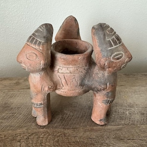 May include: A terracotta vessel featuring a central cylindrical opening supported by three stylized figures. The figures have carved facial features and geometric patterns in dark gray and reddish-brown clay, creating a tripod structure.