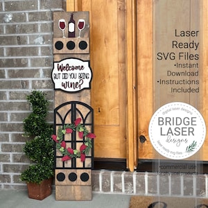 May include: A wooden sign with a welcome message that says "Welcome but did you bring wine?" The sign has a wine bottle and two wine glasses at the top, a wreath in the middle, and three black circles at the bottom. The sign is in front of a brick wall and a wooden door.