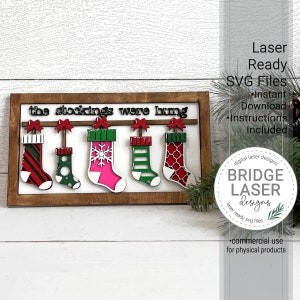 May include: A wooden sign with the text "the stockings were hung" and five colorful stockings hanging from a line. The sign is framed in wood and has a white background. The stockings are red, green, pink, and white with various patterns.