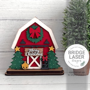 May include: A layered wooden barn-shaped decoration featuring a red facade, a green wreath with a red bow, two green Christmas trees, a yellow star on each side, a yellow bell, a black lantern, and a sign with the text Merry Christmas.
