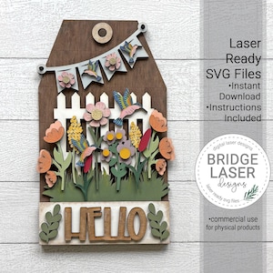 May include: Wooden laser-cut wall art featuring a garden scene with flowers, hummingbirds, and the word "HELLO." The design includes a white picket fence, a banner, and a brown tag shape. The text "Laser Ready SVG Files" is also visible.