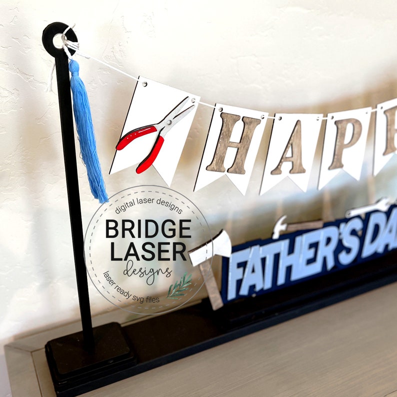 May include: A wooden Father's Day banner with the words "HAPPY FATHER'S DAY" in a mix of white, brown, and blue. The banner includes images of tools like pliers and an axe. A blue tassel hangs from the banner's support.