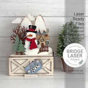 May include: A layered wooden winter scene display featuring a snowman with a black top hat and red scarf, a small house, pine trees, and snow-capped mountains. A blue tag attached to the base features the text Let it SNOW with snowflake accents.