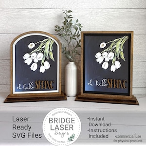 May include: Two wooden signs with a dark background, featuring white tulip illustrations and the text "oh hello SPRING". One sign is arched, the other rectangular. The signs are on wooden stands. A white vase with greenery is between them.