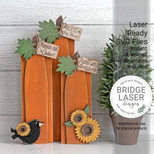 May include: Three orange wooden pumpkin cutouts with green leaves and small rectangular tags reading Bless This Home, Pumpkins and Cider, and Gather with Gratitude. A black bird with a sunflower and two additional sunflowers are attached.