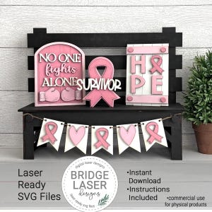Peut inclure: Un banc en bois noir pr&eacute;sente des d&eacute;corations roses et blanches d&eacute;coup&eacute;es au laser. Les d&eacute;corations comprennent une enseigne avec les mots "NO ONE FIGHTS ALONE", un ruban rose et une enseigne avec le mot "HOPE". Une banni&egrave;re avec des rubans et des c&oelig;urs roses est suspendue en dessous.