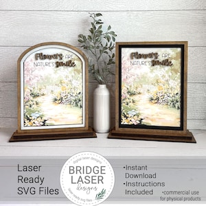 May include: Two wooden framed signs with the text "Flowers are Nature's Smile" above a watercolor image of a garden path. One sign is arched, the other rectangular. The signs are on a wooden base. A white vase with greenery is between the signs.