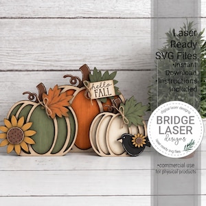 Peut inclure: Une décoration en bois superposée représentant trois citrouilles verte, orange et crème, agrémentée d'un tournesol, de feuilles d'automne, d'un petit oiseau noir et d'un panneau rectangulaire avec le texte hello FALL, attaché avec de la ficelle.