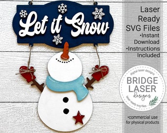 May include: A wooden snowman decoration with a navy blue sign that reads "Let it Snow". The snowman has a light blue scarf, red mittens, and a red star. The sign includes snowflakes. The image also includes the text "Laser Ready SVG Files".