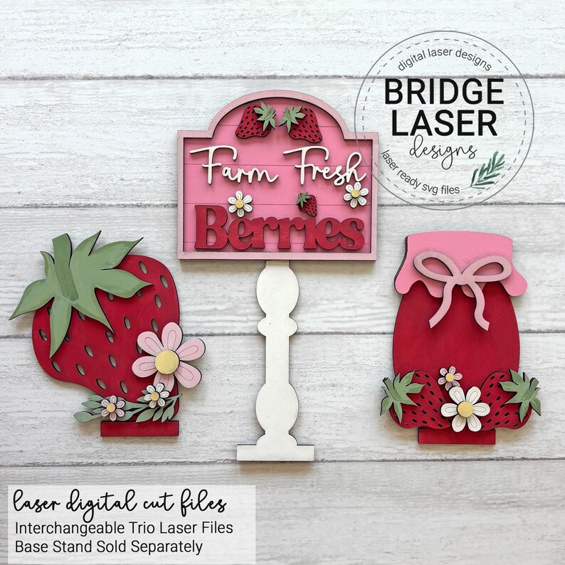 May include: Wooden laser-cut trio of strawberry-themed decorations. Includes a sign that reads "Farm Fresh Berries", a large strawberry, and a jar of jam. The sign and jar have pink accents, and the strawberry has green leaves.